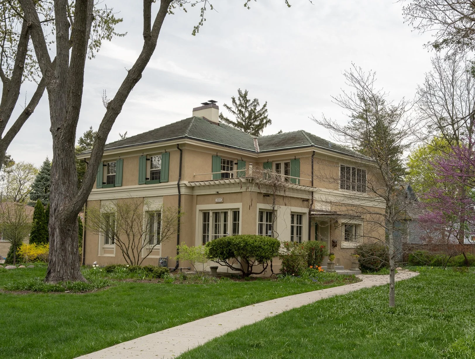 Two-story beige house with green shutters, front porch, curved sidewalk, large trees and a green lawn under an overcast sky.