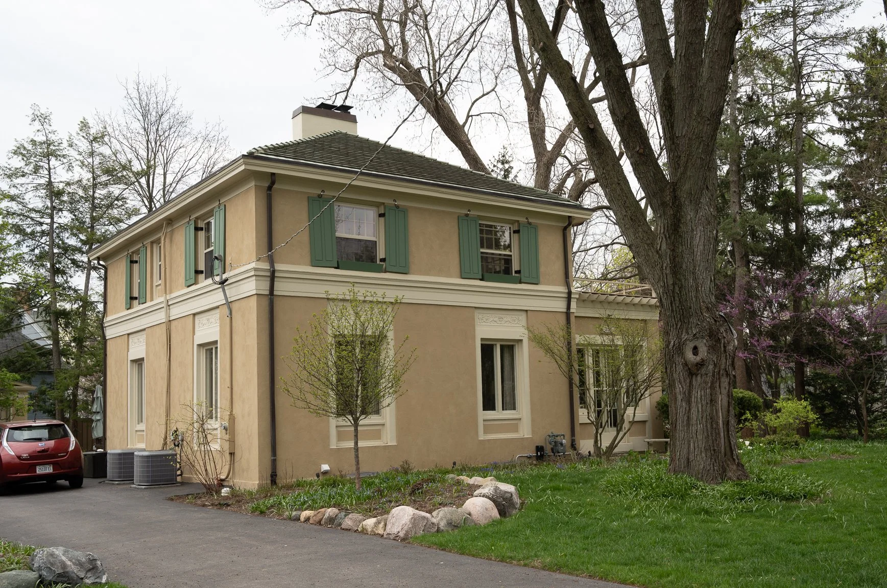 Two-story beige stucco house with green shutters, large tree in front and a driveway with a red car.