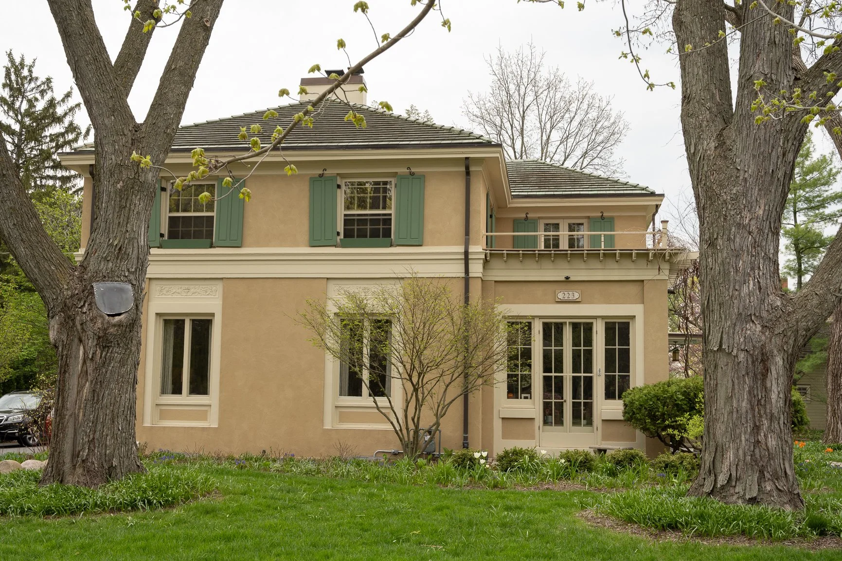Two-story beige stucco house with green shutters and French doors, framed by two large trees and a green lawn with spring foliage.