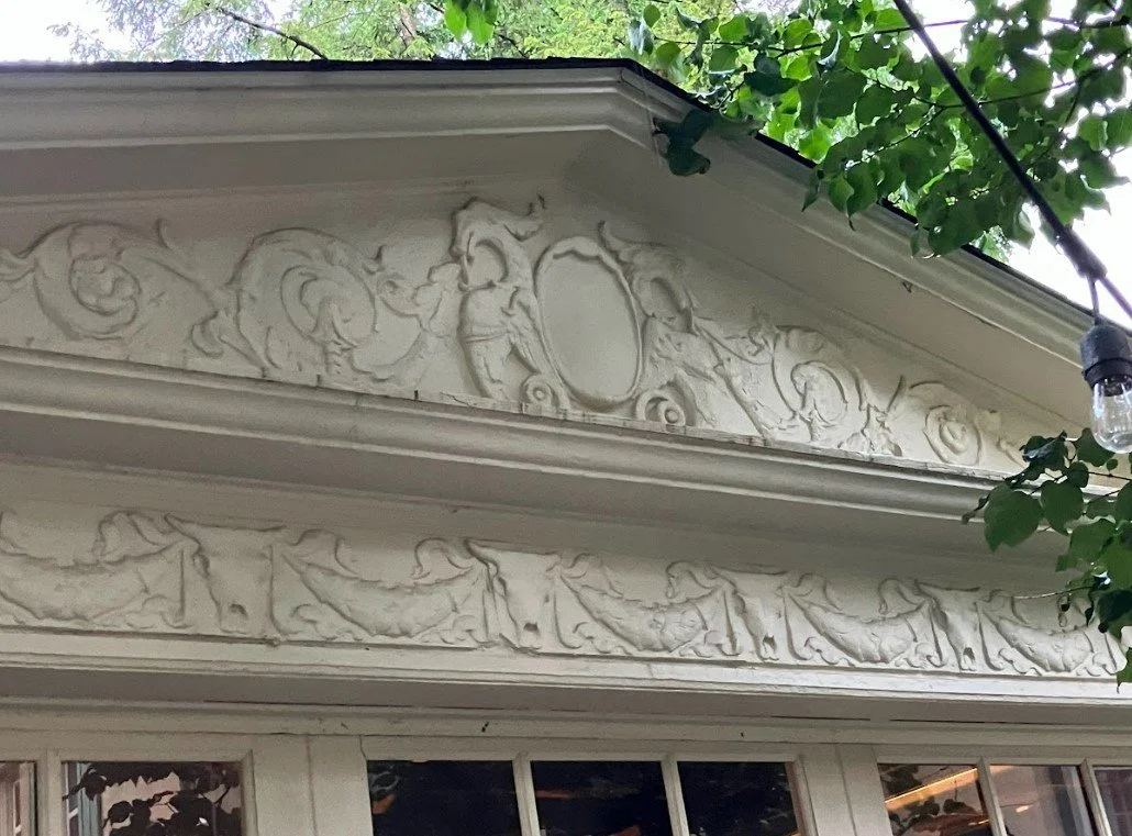White ornate plaster pediment with carved scrolls and floral motifs above a window, framed by green leaves and a hanging light bulb.