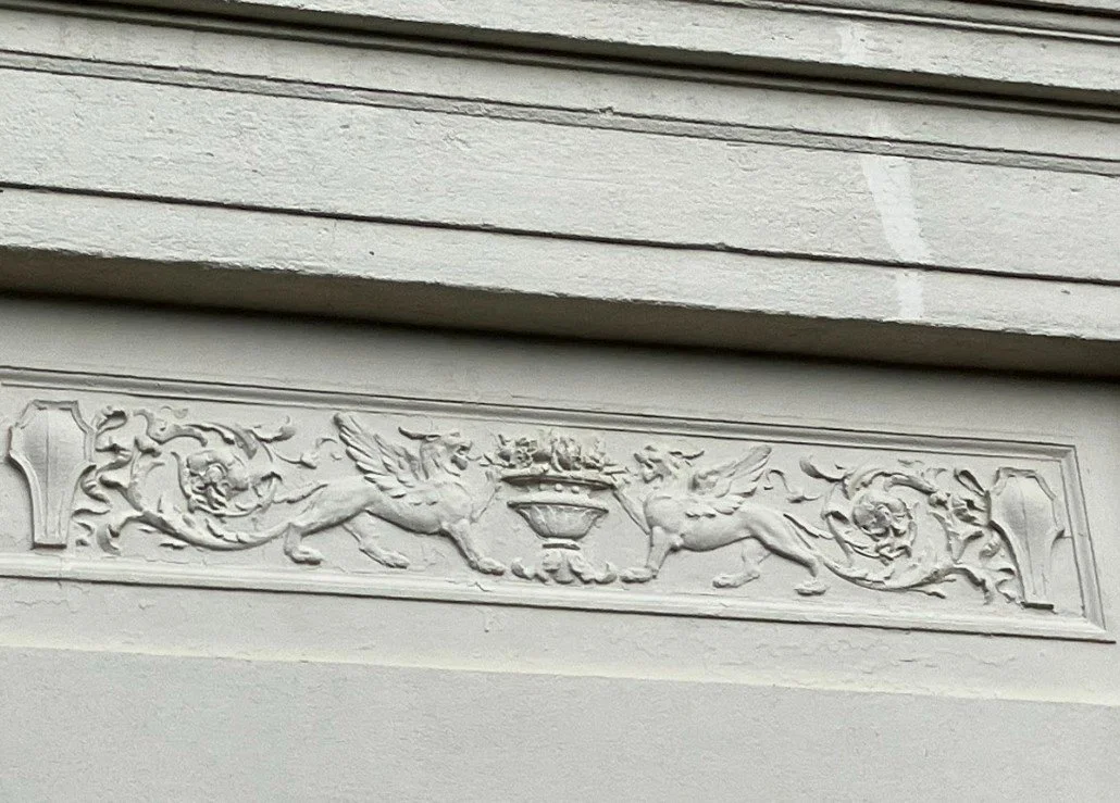 Ornate plaster frieze showing two winged griffins facing a central urn filled with flowers, flanked by scrolling floral motifs.