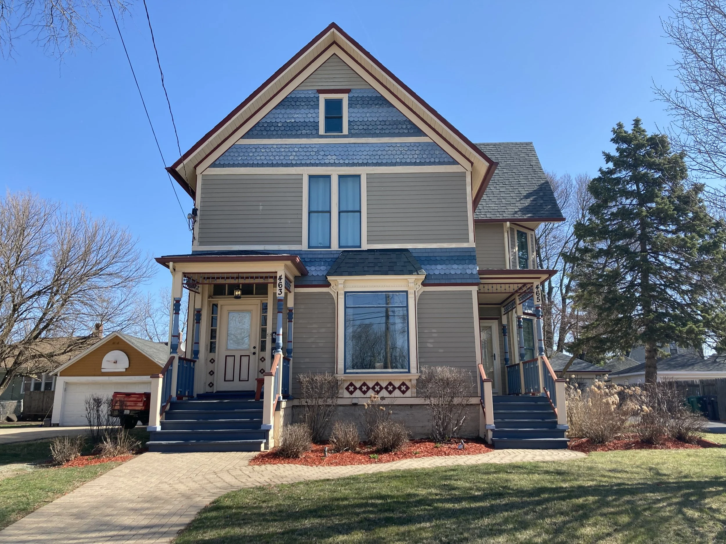 Two-story Victorian-style house with blue-gray siding, two front porches, a pathway, detached garage, and clear blue sky.