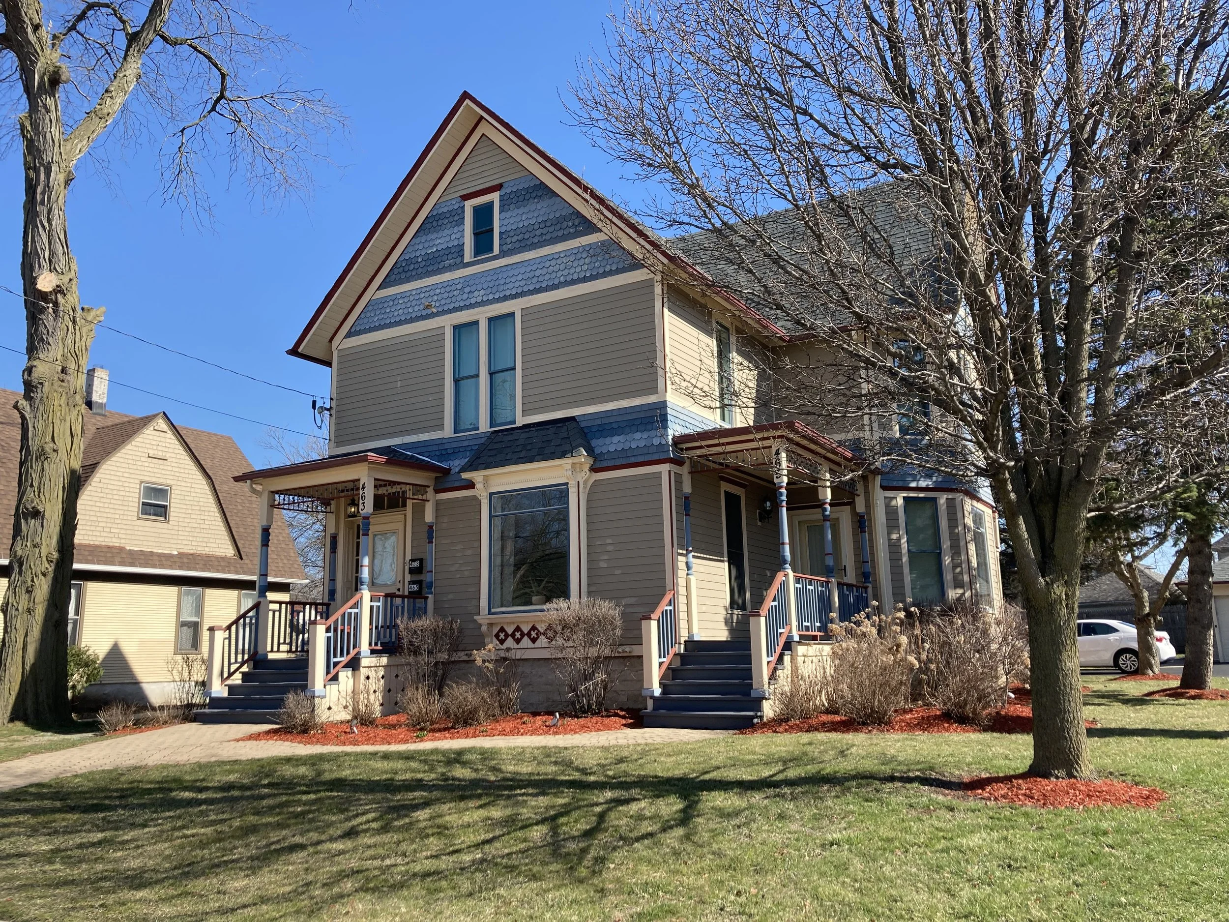 Two-story Victorian-style house with blue trim and porches, leafless trees in the yard, and a sunny green lawn.