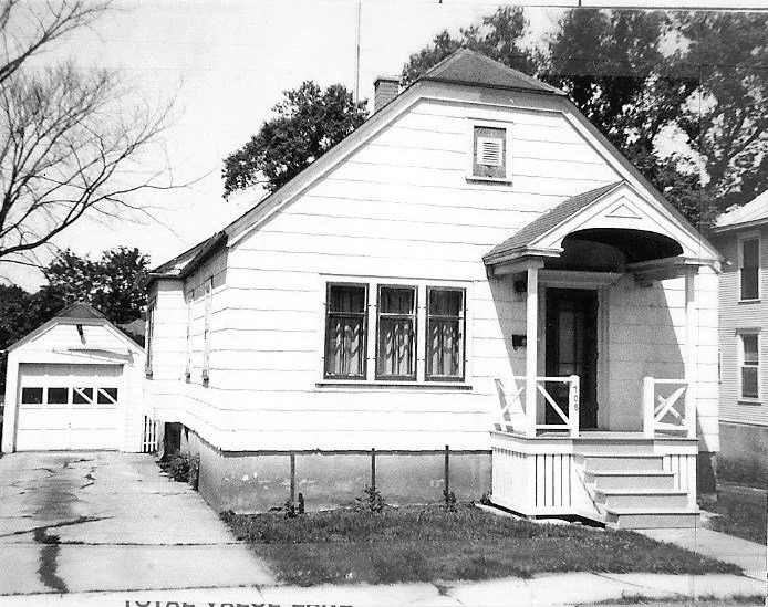Black-and-white photo of a small white single-story house with a covered front porch, steps, driveway leading to a detached garage, and trees in the background.