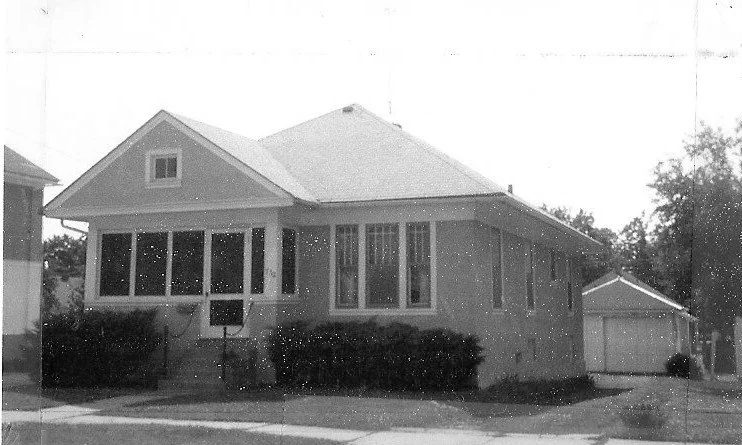 Black-and-white photo of a single-story bungalow with a screened front porch, steps and shrubs at the front, and a detached garage to the right.