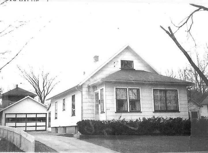 Black-and-white photo of a single-story bungalow with a detached two-car garage, front driveway, low hedges, and leafless trees.