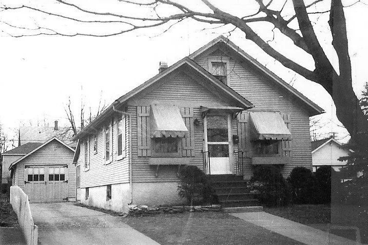 Black-and-white photo of a small bungalow with front steps, awnings over windows, a detached garage at the end of a driveway, and leafless trees framing the yard.