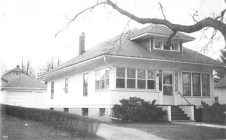Black-and-white photo of a single-story bungalow with an enclosed front porch, small front steps, driveway leading to a detached garage, and bare tree branches overhead.