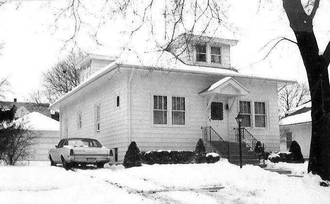 Black-and-white photo of a single-story house with a dormer, snow-covered yard, front steps and a parked car.