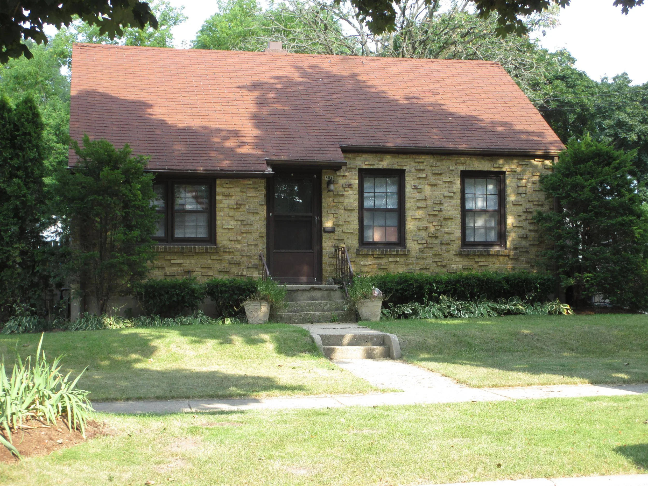 Small yellow-brick house with a steep red shingled roof, central dark front door, three front windows, concrete steps and shrubs on a green lawn.