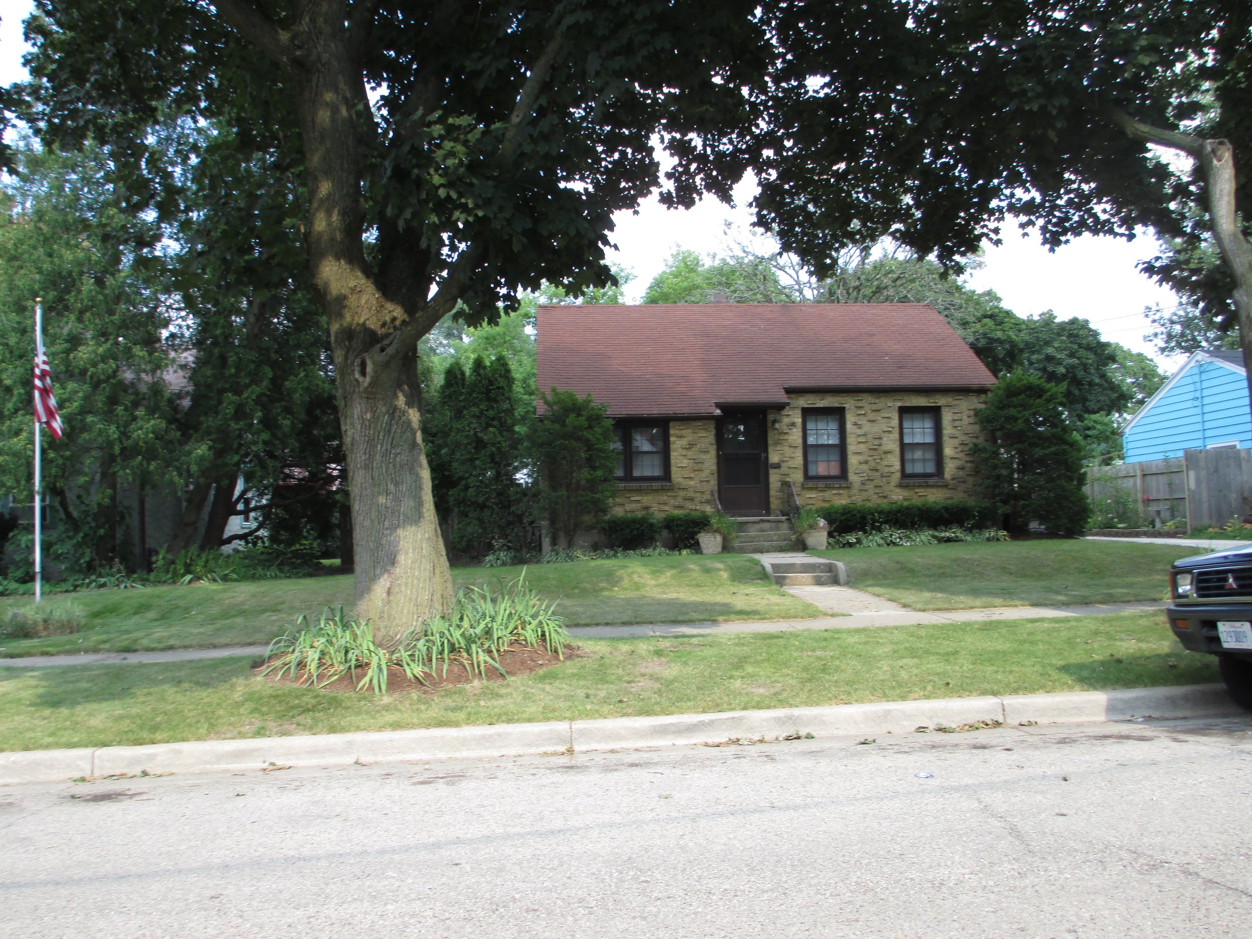 Small brick house with a red roof partly shaded by a large tree, front lawn, flagpole on the left and a parked truck at right.