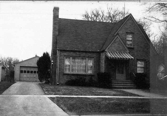 Black-and-white photo of a brick Tudor-style house with a steep gable and striped awning over the front door, a driveway leading to a detached garage on the left, and leafless trees in the background.