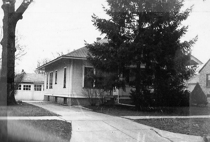 Black-and-white photo of a single-story bungalow with a driveway, large evergreen tree, and detached garage.