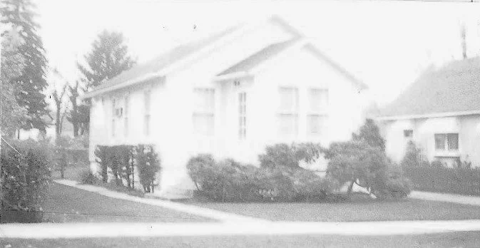 Black-and-white photo of a small single-story bungalow with shrubs along the front and a sidewalk in the foreground.