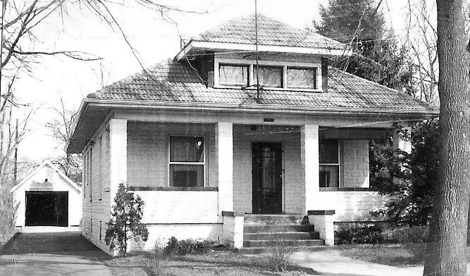 Black-and-white photo of a single-story bungalow with a covered front porch supported by square columns, central steps, and a detached garage visible down the driveway at left