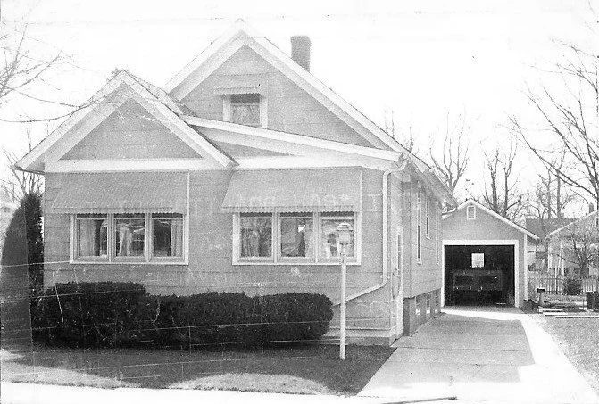 Black-and-white photo of a one-story house with front awnings, shrubs, driveway leading to a detached garage.