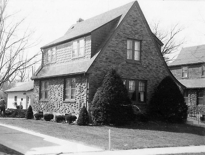 Black-and-white photo of a two-story brick house with a steep gabled roof, front shrubs and a sidewalk.