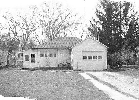 Small single-story house with an attached one-car garage, a bicycle leaning against the front wall, leafless trees behind it, and a driveway leading up to the garage.