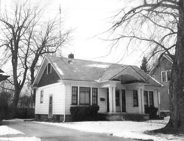 Black-and-white photo of a single-story bungalow with a covered front porch, leafless trees, and light snow on the ground.
