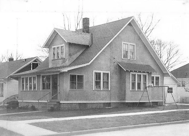 Black-and-white photo of a two-story bungalow with a front porch, dormer window, chimney, and a small swing set on a corner lot.