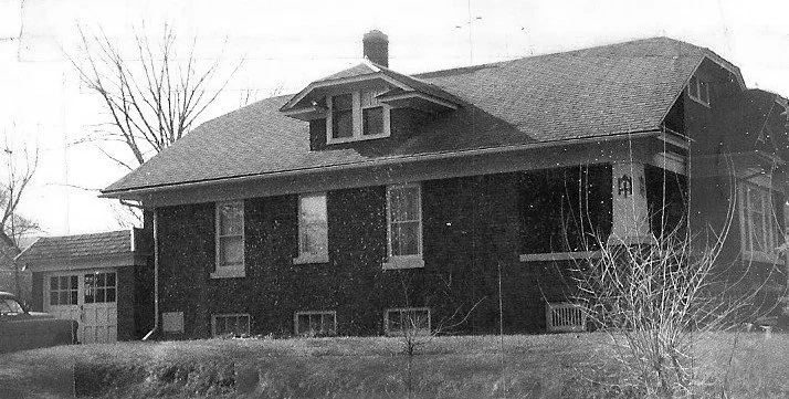 Black-and-white photo of a one-story brick bungalow with a central dormer and chimney, small front yard, detached garage and a parked car.