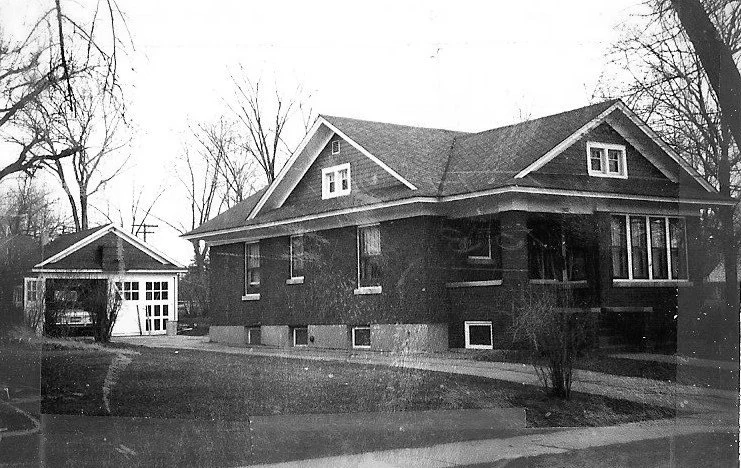Black-and-white photo of a single-story brick bungalow with a detached garage, bare trees, and a front lawn.