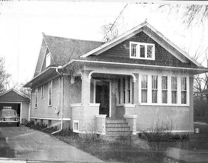 Black-and-white photo of a Craftsman bungalow with a covered front porch, steps leading up, a detached garage, and a vintage car parked in the driveway.