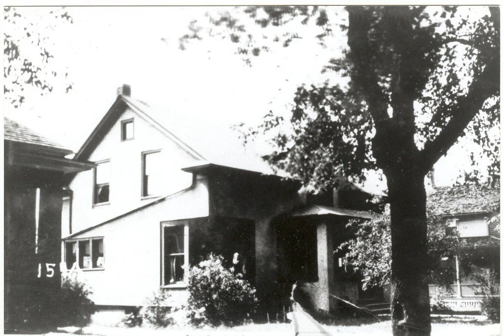 Black-and-white photo of a two-story house with a covered front porch, large tree in the yard, and a neighboring house visible at right.