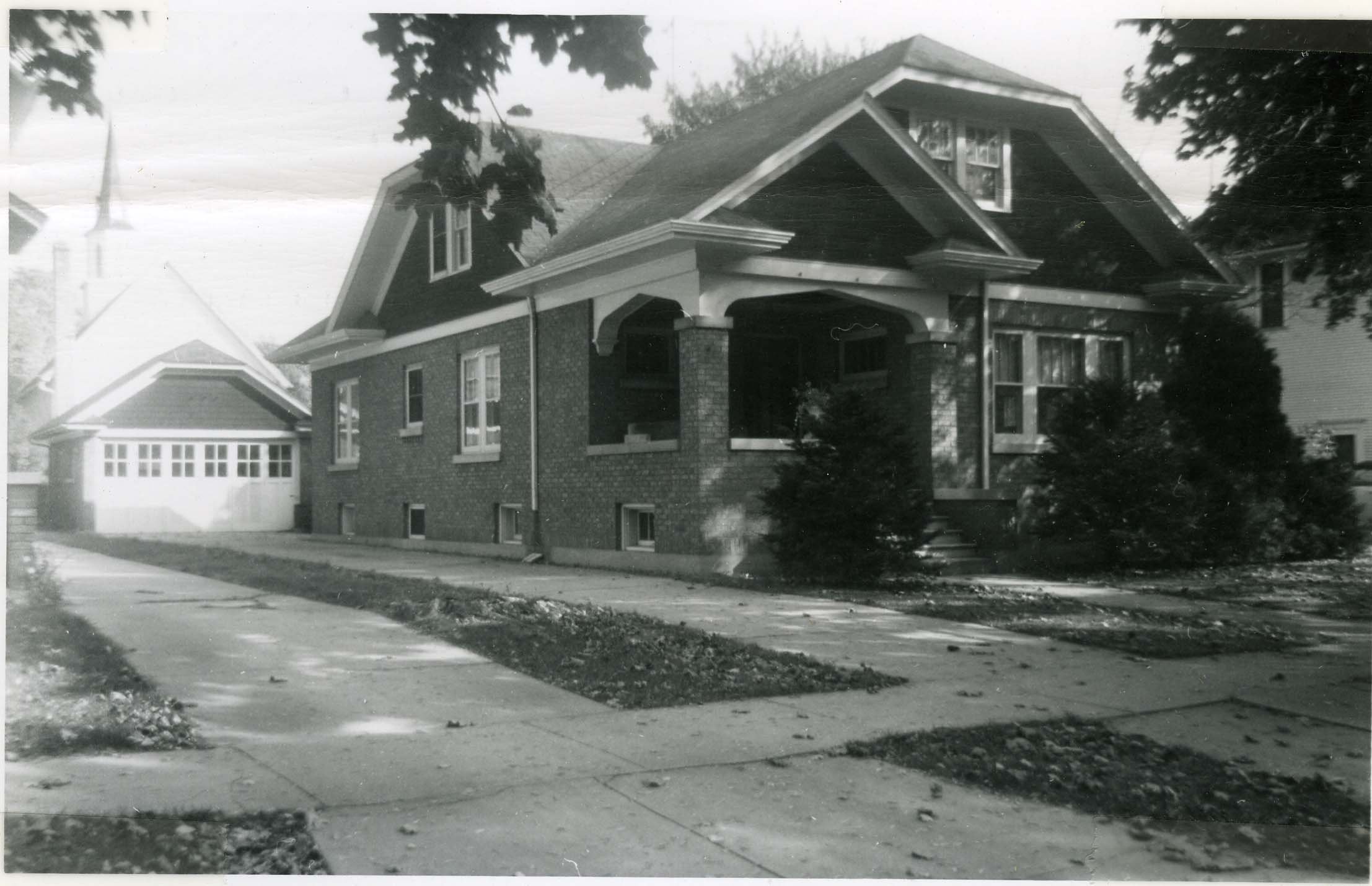 Black-and-white photo of a brick bungalow with a covered front porch, dormer windows, detached garage, and a tree-lined sidewalk.