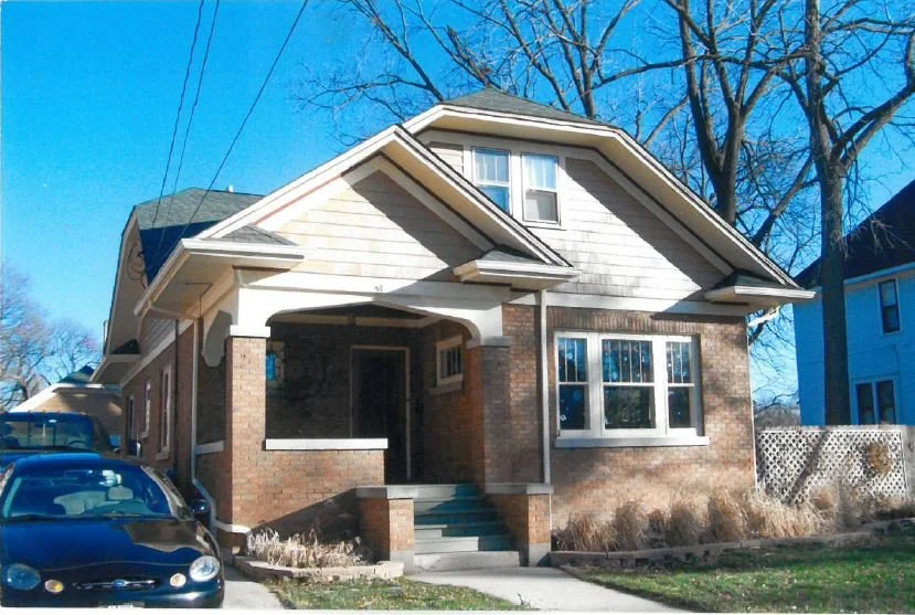 Brick bungalow-style house with a covered front porch and steps, parked cars in the driveway, leafless trees behind it and a clear blue sky.