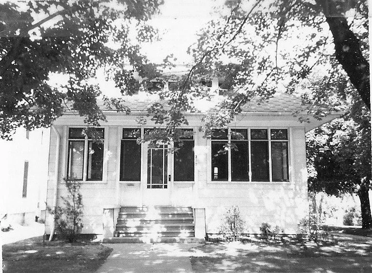 Black-and-white photo of a small single-story house with an enclosed front porch, six steps up to a central door, and large trees casting shadows.