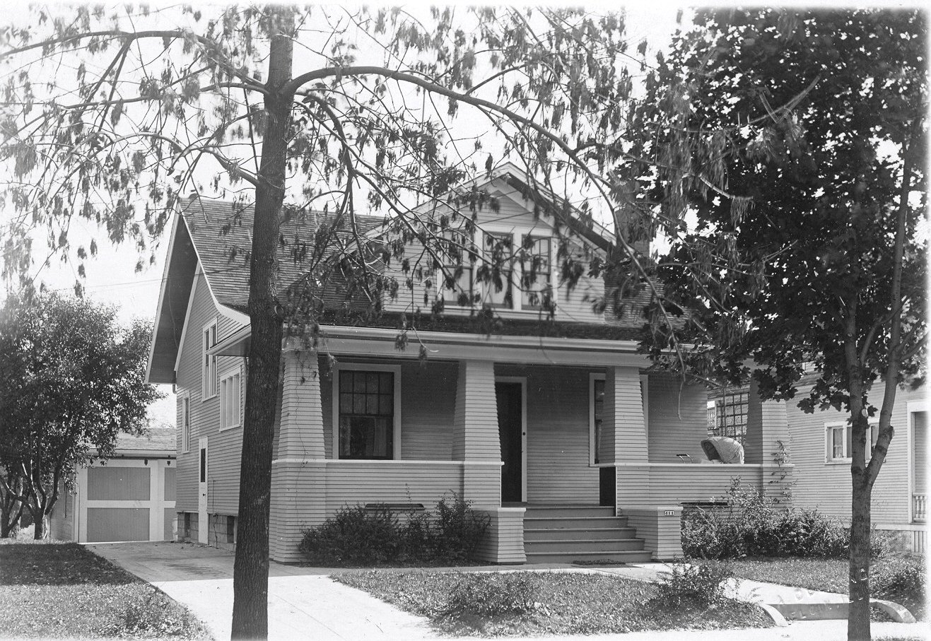 Black-and-white photo of a two-story bungalow with a wide front porch and steps, trees in the yard, and a detached garage to the left.