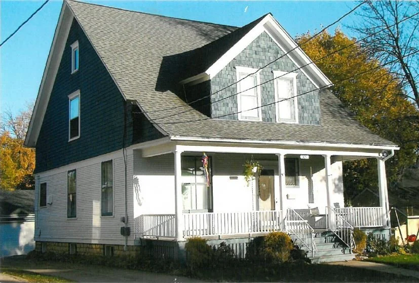 Front two-story white house with a covered front porch, steep gray gabled roof, and hanging plants, set on a tree-lined street.