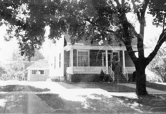 Black-and-white photo of a small house with a covered front porch and steps, a large tree in the front yard, and a detached garage to the left.