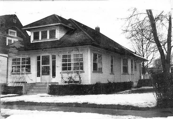 Black-and-white photo of a single-story bungalow-style house with a wide front porch, multiple windows, a dormer roof, snow on the lawn and a leafless tree at the right.