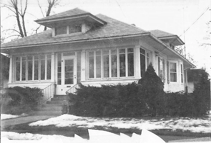 Black-and-white photo of a single-story bungalow-style house with an enclosed front porch, steps leading to the central door, shrubs along the front, and patches of snow on the lawn.