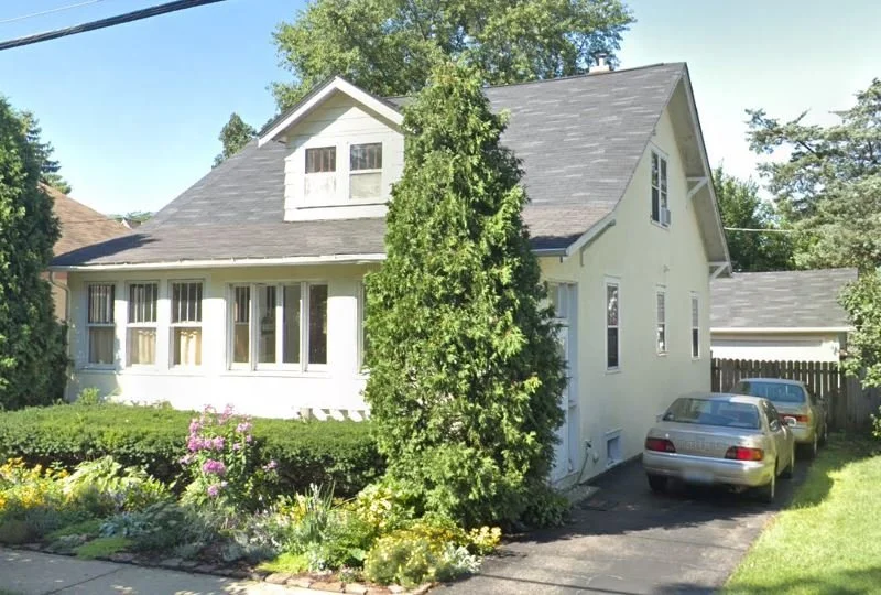Light-colored bungalow with enclosed front porch, tall evergreen and flowerbed in front, and a driveway on the right with two parked cars.