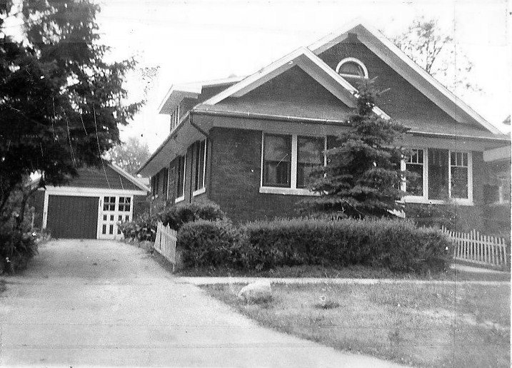 Black-and-white photo of a brick bungalow with shrubs and a small pine in front, a white picket fence, driveway, and detached garage.