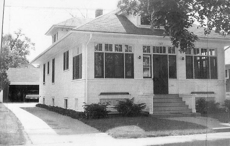 Black-and-white photo of a two-story bungalow with enclosed front porch, steps up to a central door (number 707), driveway leading to a detached garage.