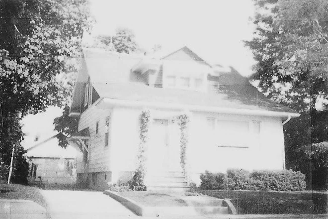Black-and-white photo of a two-story suburban house with front steps and an ivy-framed doorway, flanked by trees and shrubs.