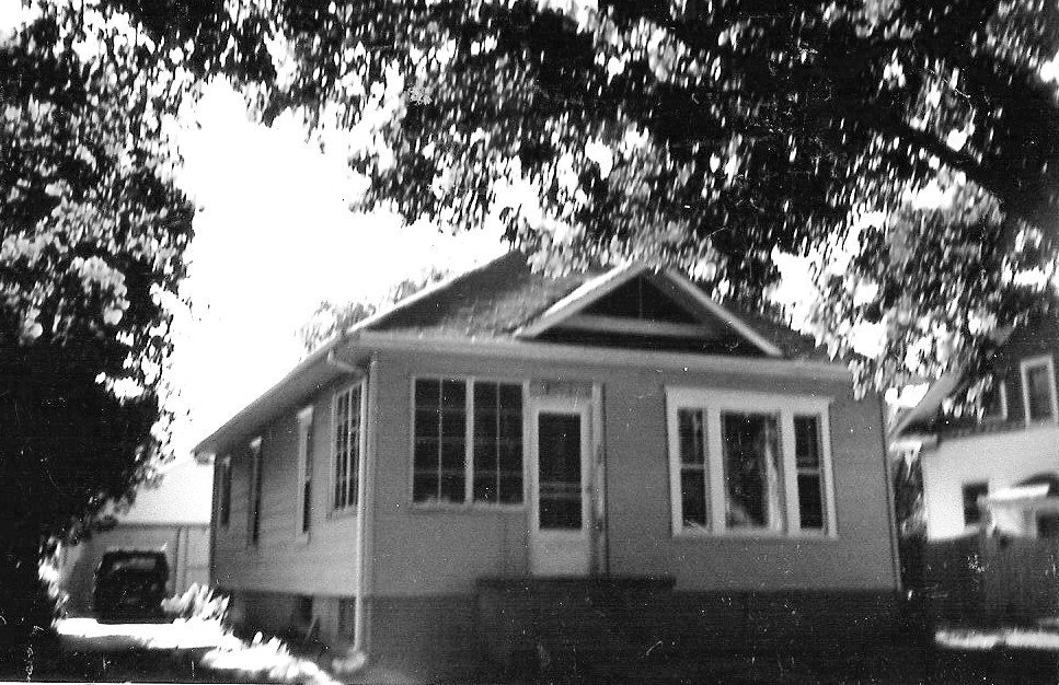 Black-and-white photo of a small single-story house with a front porch door and windows, large trees overhead and a car parked in the driveway to the left.