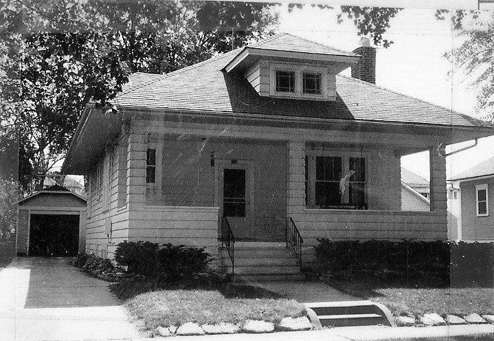 Black-and-white photo of a single-story bungalow with a front porch, steps, shrubs, and a detached garage at the end of a driveway.