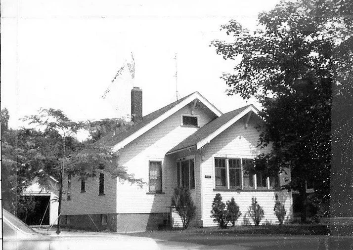 Black-and-white photo of a small single-story Craftsman-style house with a front porch, shrubs, and trees.