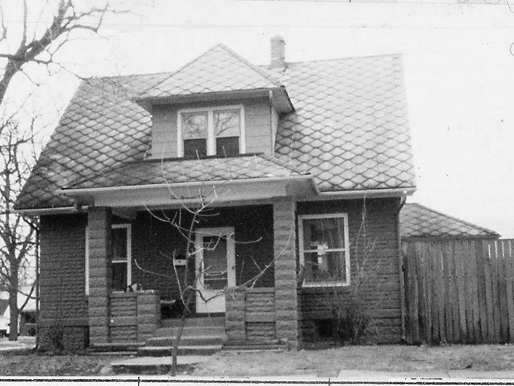Black-and-white photo of an older two-story house with a covered front porch, dormer window, patterned shingle roof, leafless tree in front, and wooden fence to the right.