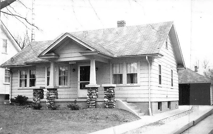 Black-and-white photo of a single-story bungalow with a small front porch supported by stone piers and a detached garage at the side.