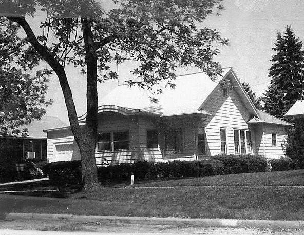 Black-and-white photo of a single-story wood-frame house with a front porch, low shrubs and a large tree in the front yard.