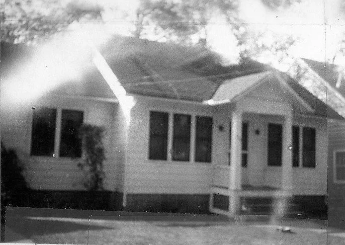 Black-and-white photo of a single-story house with a small front porch supported by columns and three front windows.