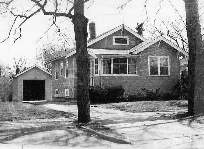 Black-and-white photo of a single-story bungalow with a front porch, driveway leading to a detached garage, and leafless trees in the yard.