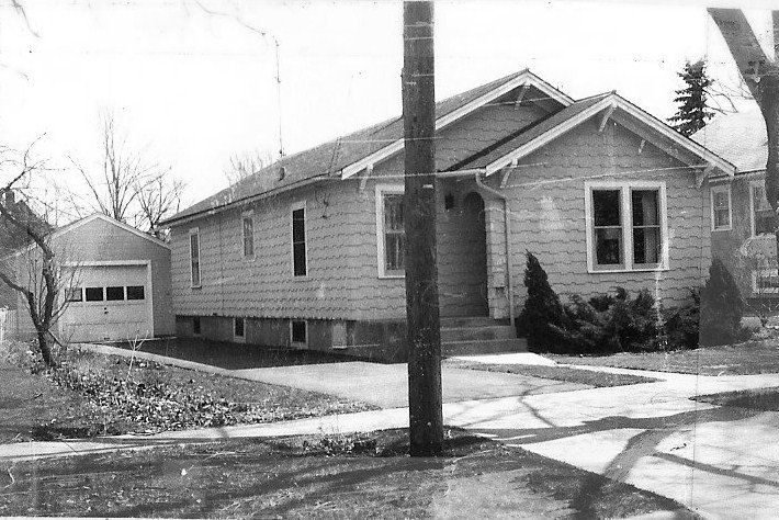 Black-and-white photo of a single-story bungalow with a detached garage, driveway, sidewalk and a utility pole in front.
