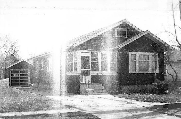 Black-and-white photo of a single-story bungalow with a small front porch and steps, detached garage to the left, driveway in front, and bare trees, with light glare across the image.